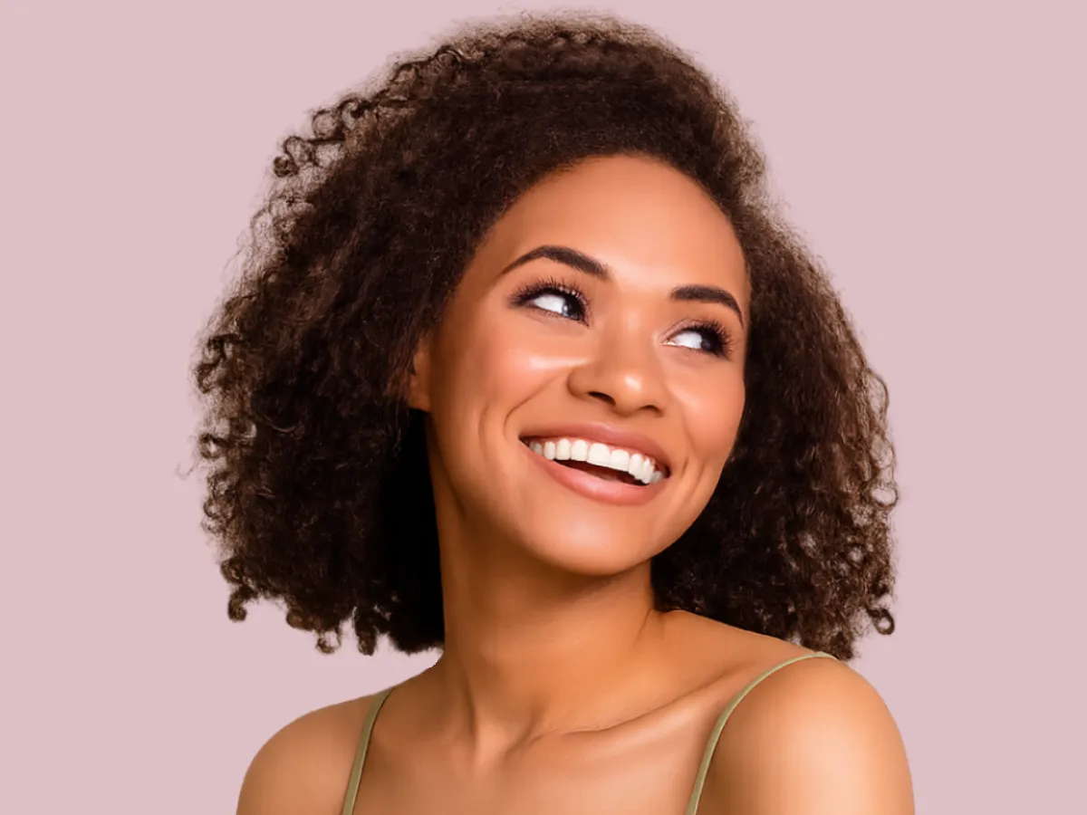 A woman with medium length curly hair stares off-camera with a big wide-mouthed smile on a light pink background